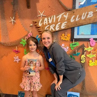 A young Cavity Free Club winner with her photo prize and a Super Smiles team member, in front of the Cavity Free Club wall, fishing-net display with the hand-painted "CAVITY FREE CLUB" sign